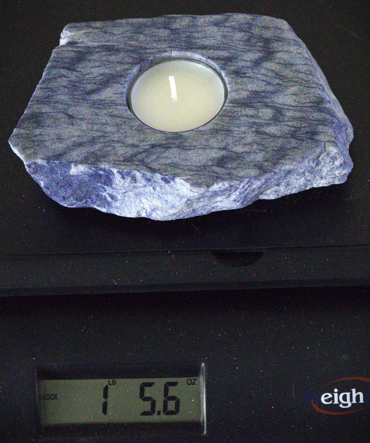 A square slate candle holder with a circular inset holding a white tea light candle, shown resting on a digital scale displaying 1.56 oz.