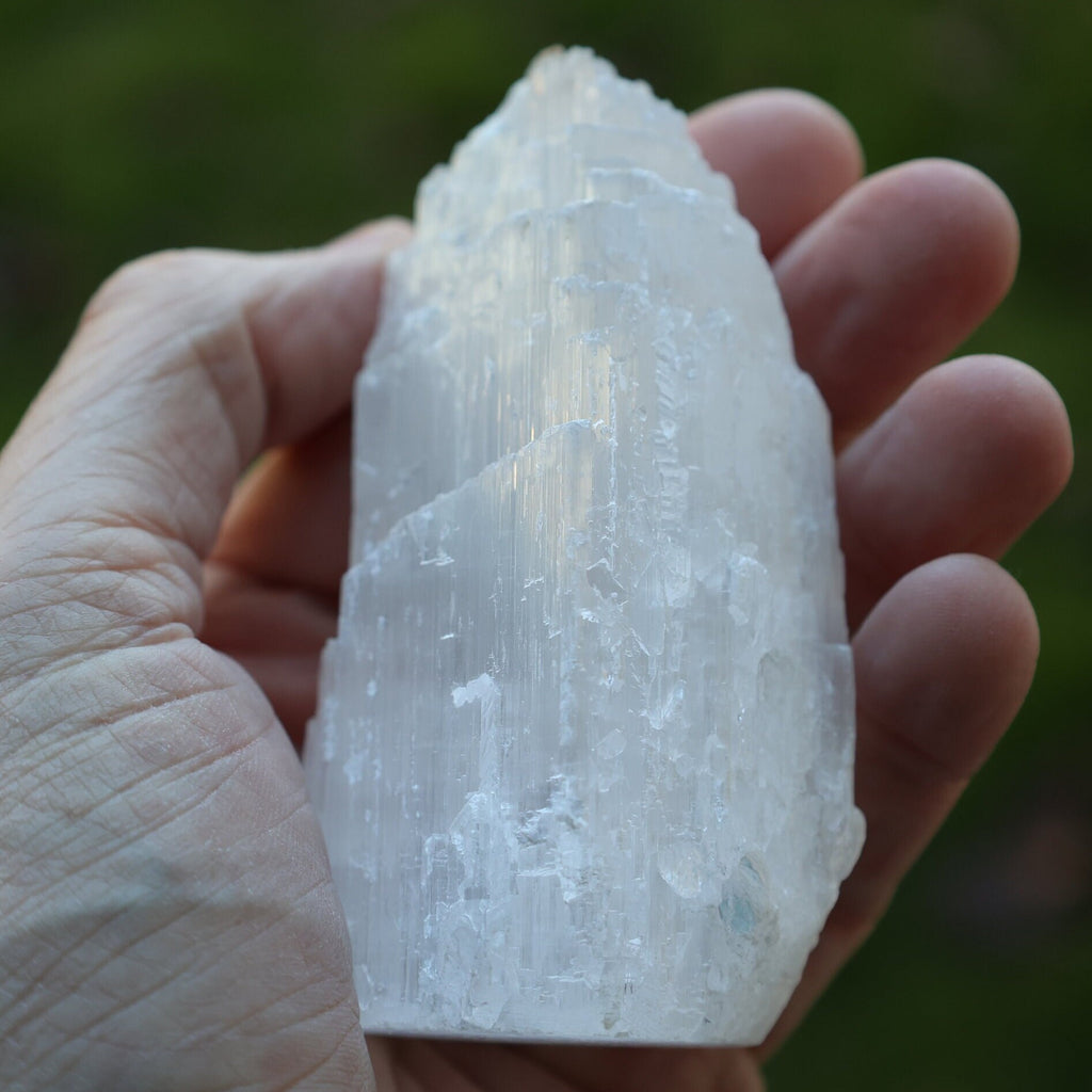 A hand holding a large, rough, translucent white crystal or mineral specimen against a blurred green outdoor background.