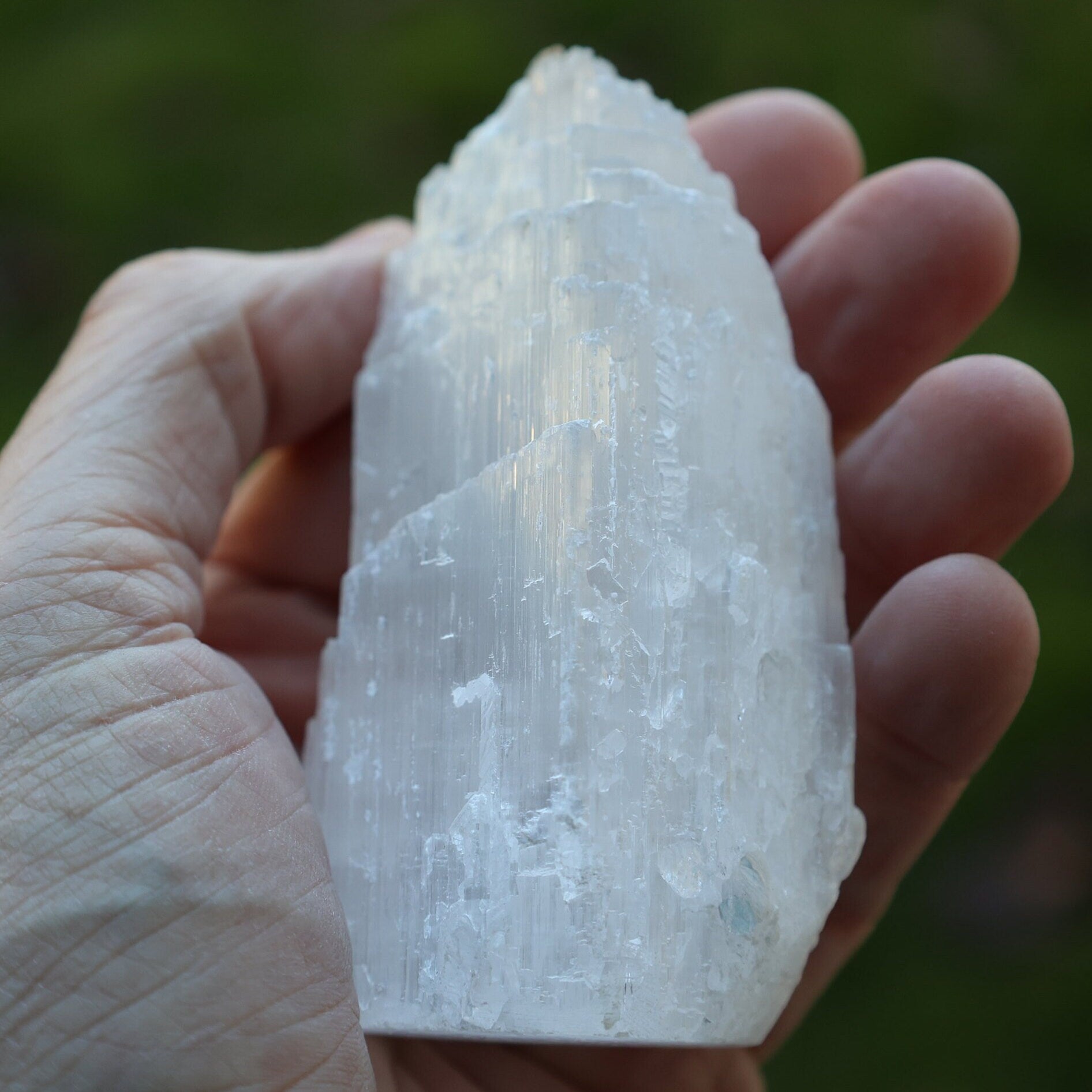 A hand holding a large, rough, translucent white crystal or mineral specimen against a blurred green outdoor background.