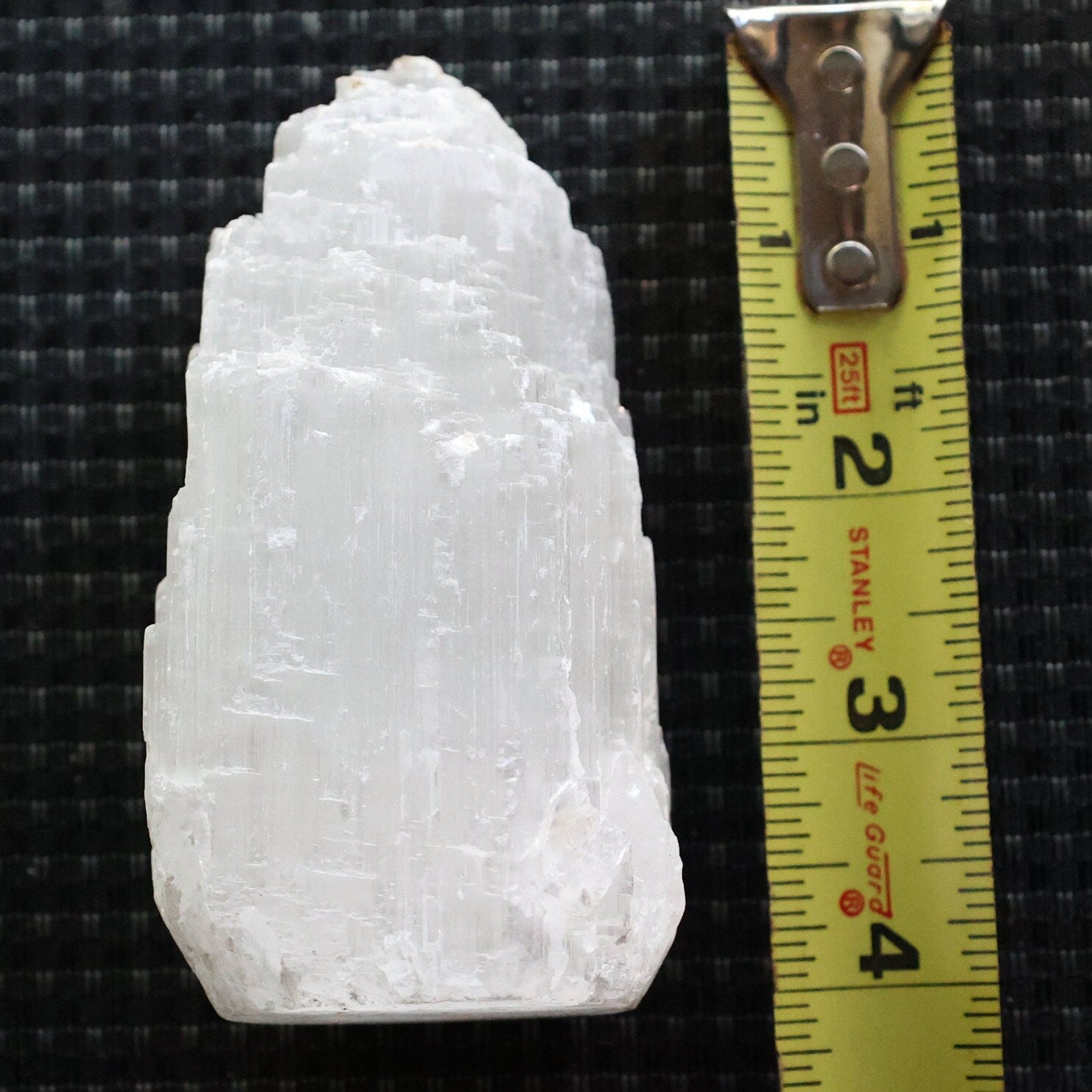 A white, textured mineral or crystal specimen shown next to a yellow Stanley tape measure on a black woven background.