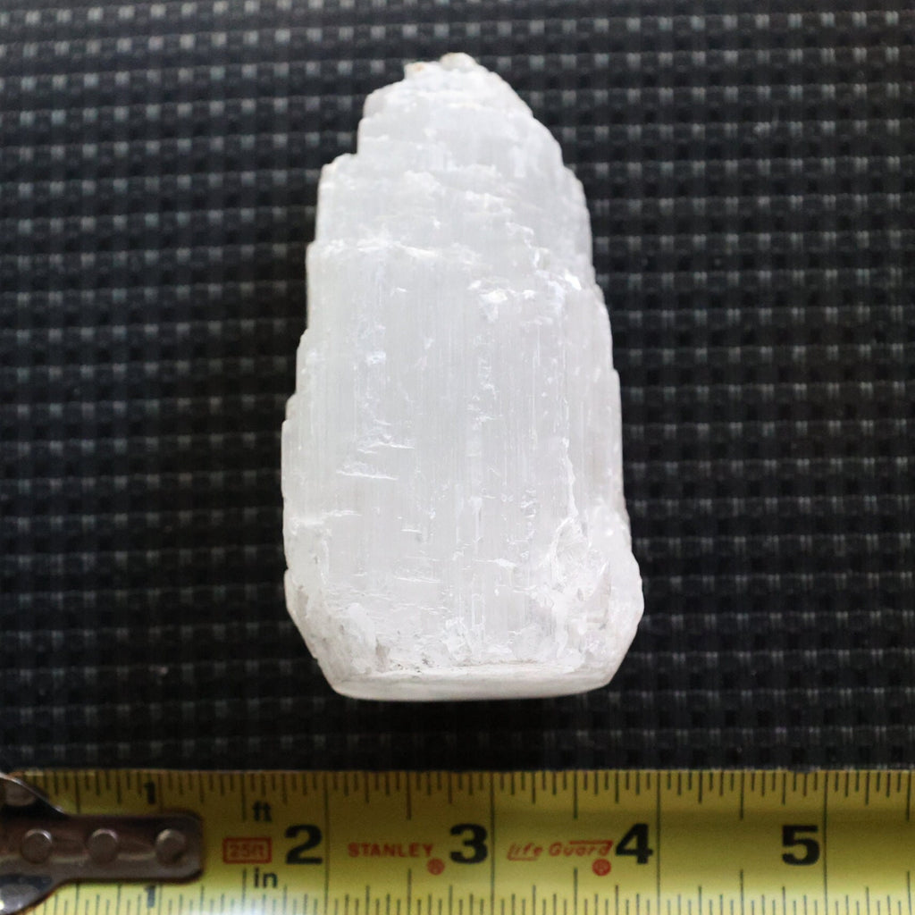 A raw, rough white crystal or mineral specimen with natural striations, shown on a black textured background with a yellow tape measure at the bottom for scale.