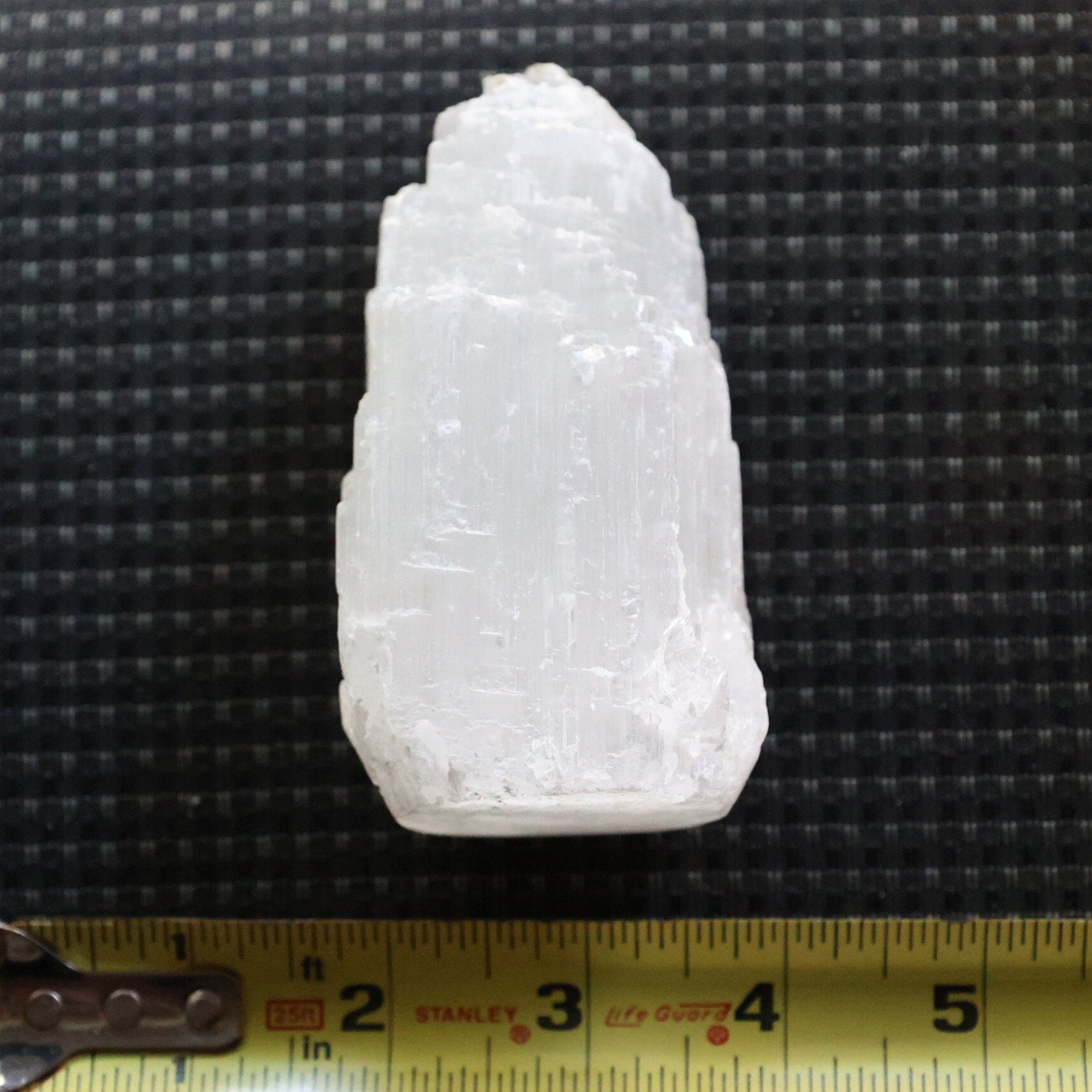 A raw, rough white crystal or mineral specimen with natural striations, shown on a black textured background with a yellow tape measure at the bottom for scale.