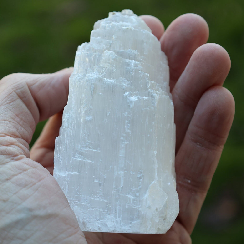 A hand holding a large, raw, pointed white crystal mineral specimen against a blurred green outdoor background.