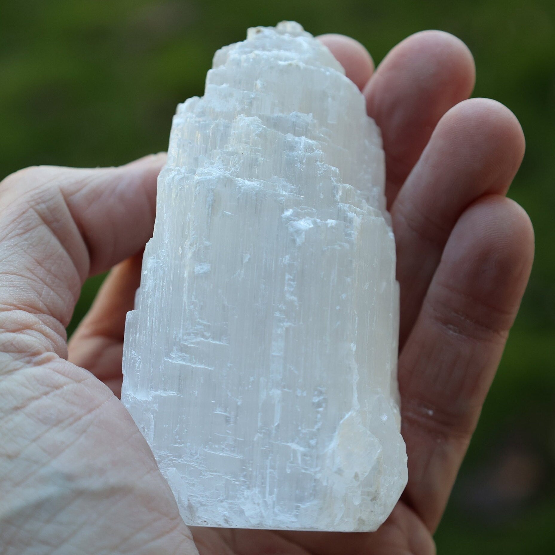 A hand holding a large, raw, pointed white crystal mineral specimen against a blurred green outdoor background.