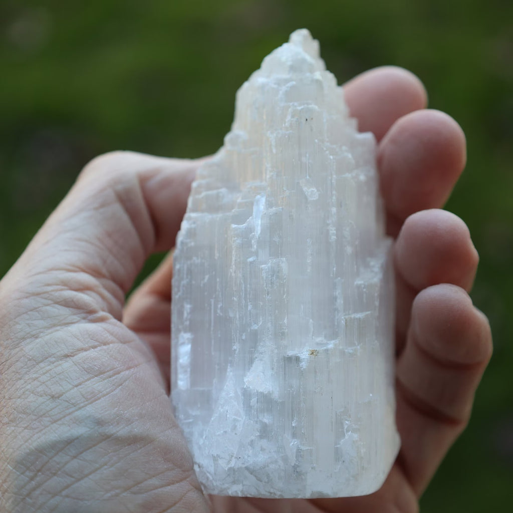 A hand holding a raw, pointed white crystal mineral specimen against a blurred green outdoor background.