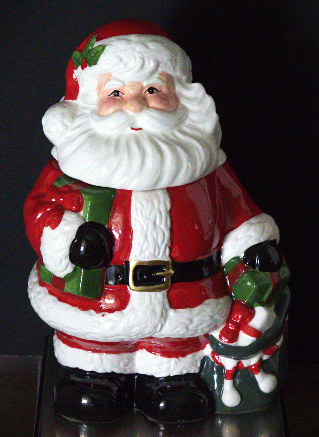 A vintage Santa Claus ceramic figurine holding a green gift box and a decorated Christmas tree, set against a dark background.