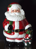 A vintage Santa Claus ceramic figurine holding a green gift box and a decorated Christmas tree, set against a dark background.