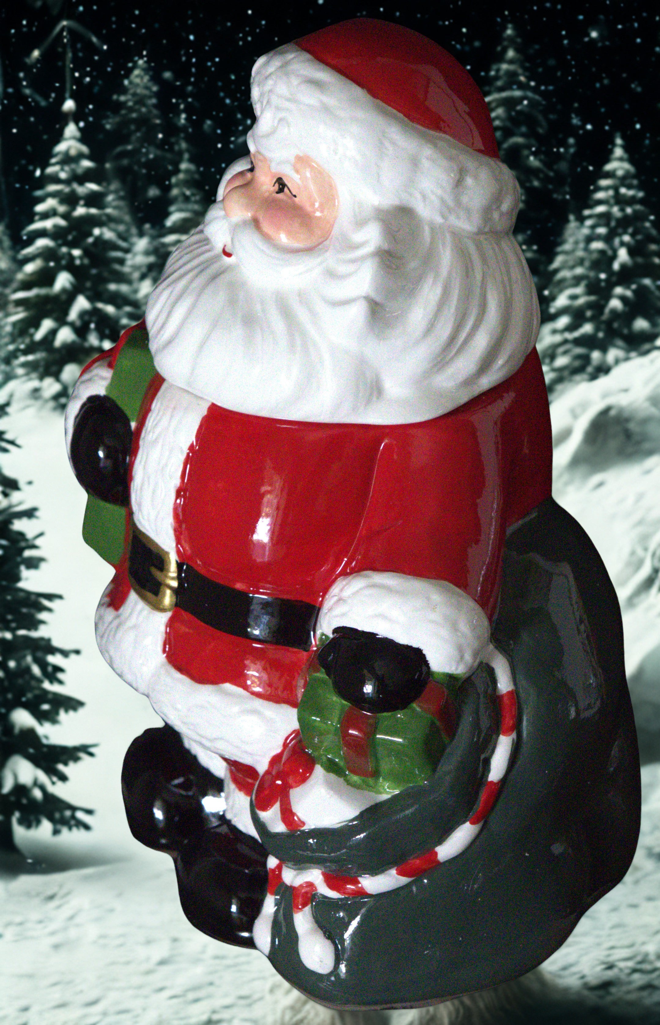 A glossy ceramic Santa Claus figurine wearing his traditional red and white suit, with a black belt, black boots, and a dark green sack on his back, holding a small green gift box. The figurine is set against a snowy night scene backdrop with pine trees.