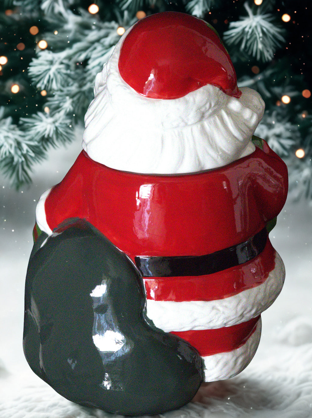 A glossy ceramic Santa Claus Cookie Jar shown from the back, wearing a red suit with white trim, a black belt, and holding a large black sack, set against a snowy outdoor holiday background with pine branches and warm lights.