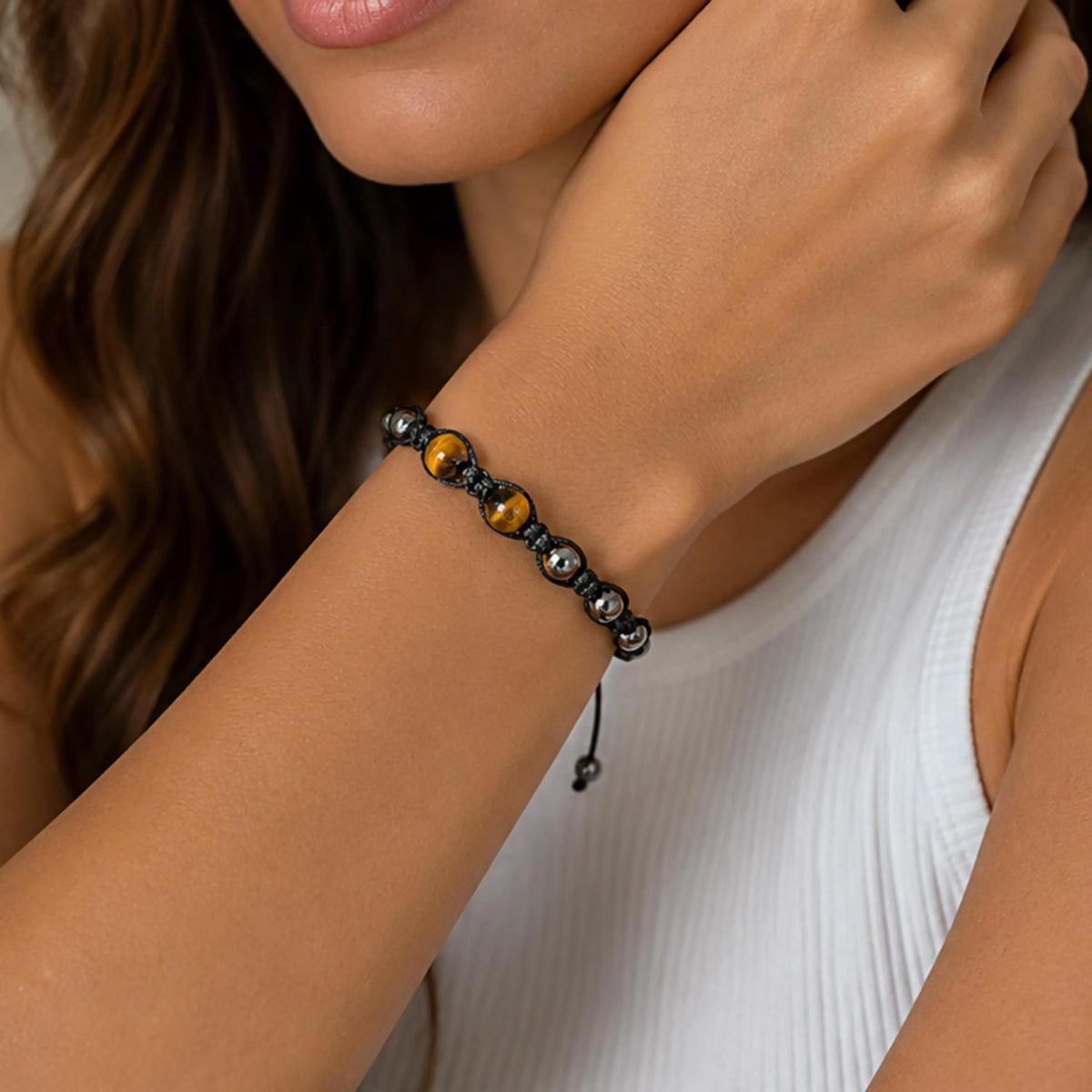 A close-up of a woman's wrist wearing a beaded bracelet with dark gray beads, two gold tiger's eye beads, and silver-toned spacer beads, paired with a white ribbed tank top.