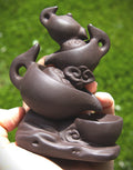 A hand holding a dark brown ceramic tea set sculptural figurine featuring a teapot, cup, and decorative cloud motifs, set against a blurred green grass background.