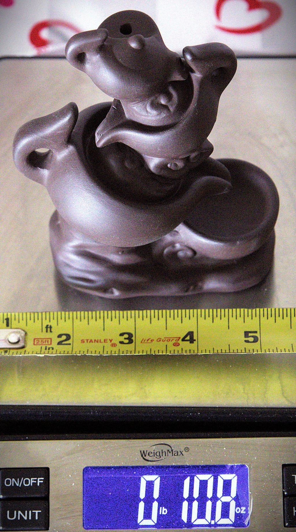 A small dark brown ceramic figurine of a stylized animal or creature, shown on top of a yellow tape measure with a digital scale below displaying weight.