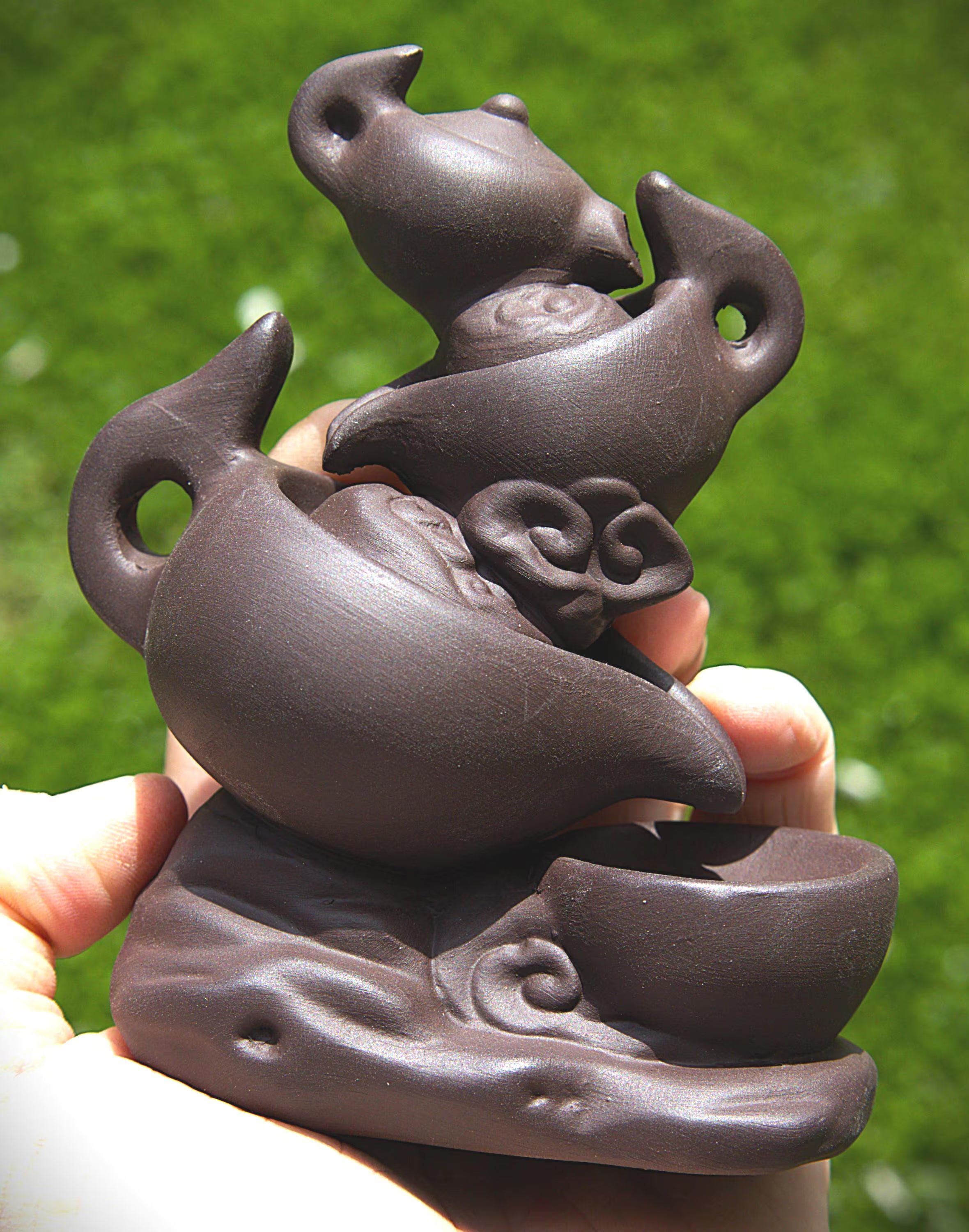A hand holding a dark brown ceramic tea set sculptural figurine featuring a teapot, cup, and decorative cloud motifs, set against a blurred green grass background.