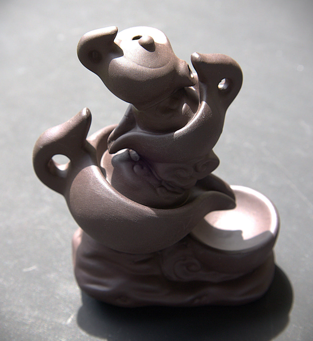 A small sculptural figurine in a matte dark brown or charcoal tone, depicting a stylized animal or creature curled around a curved form with a shallow dish-like element on the right, photographed on a dark gray surface.