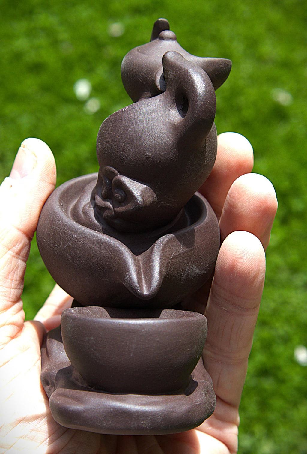 A small dark brown clay figurine of a stylized cat sitting in a bowl, held in a person's hand against a blurred green grass background.