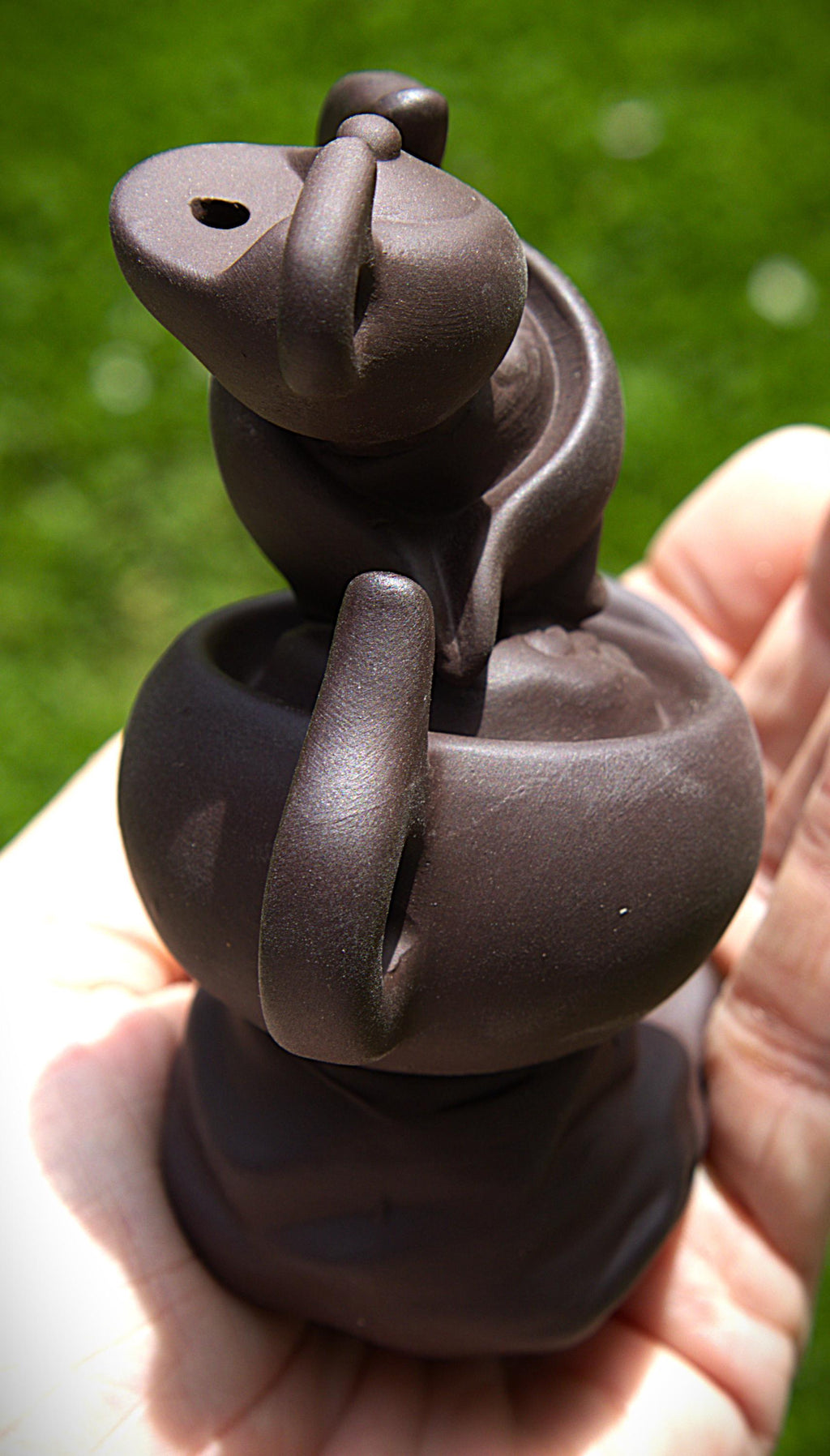 A small dark brown ceramic figurine of a stylized animal or creature, held in a person's hand against a blurred green outdoor background.