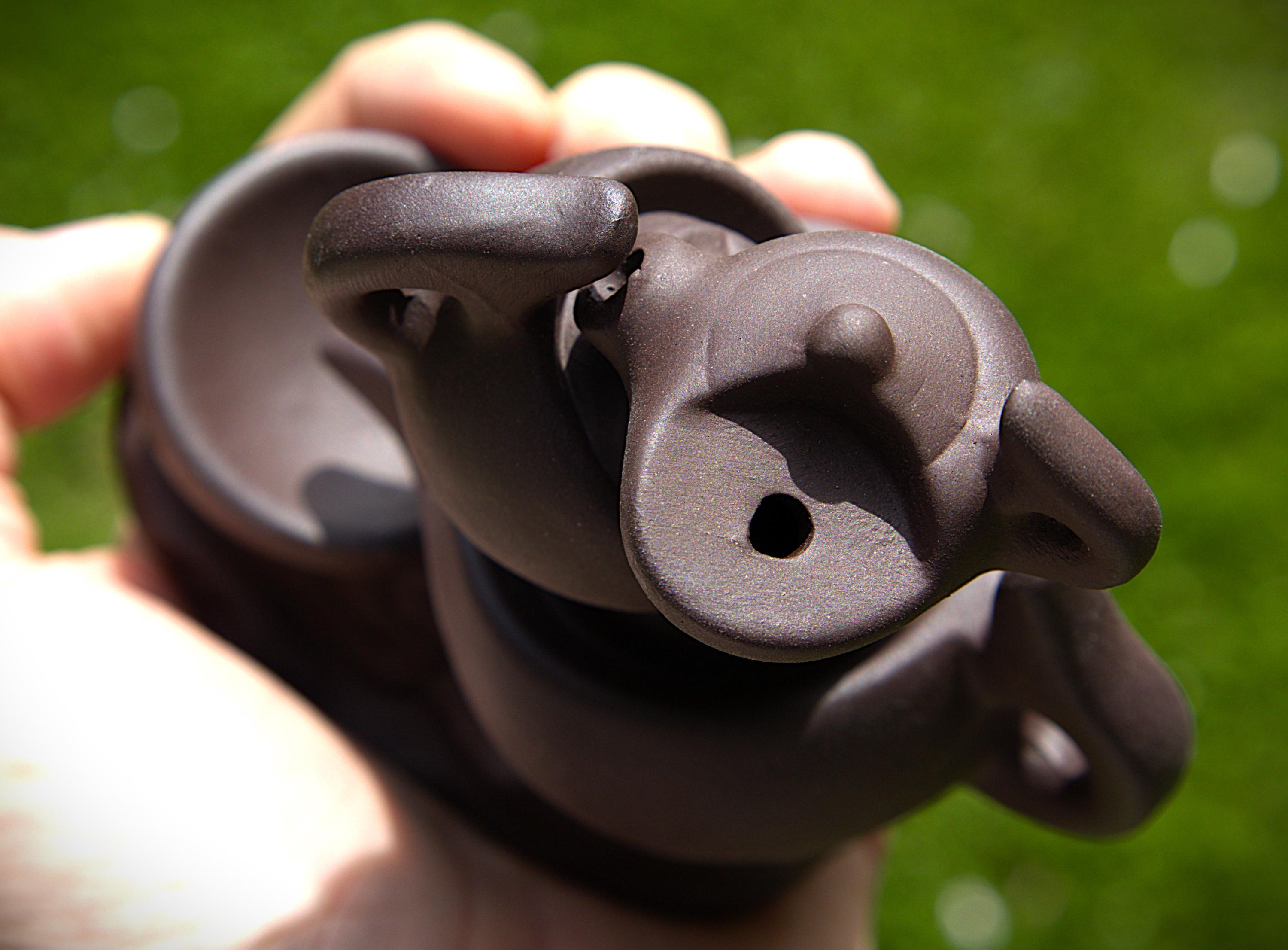 A close-up of a hand holding a dark brown ceramic teapot outdoors against a blurred green grass background.