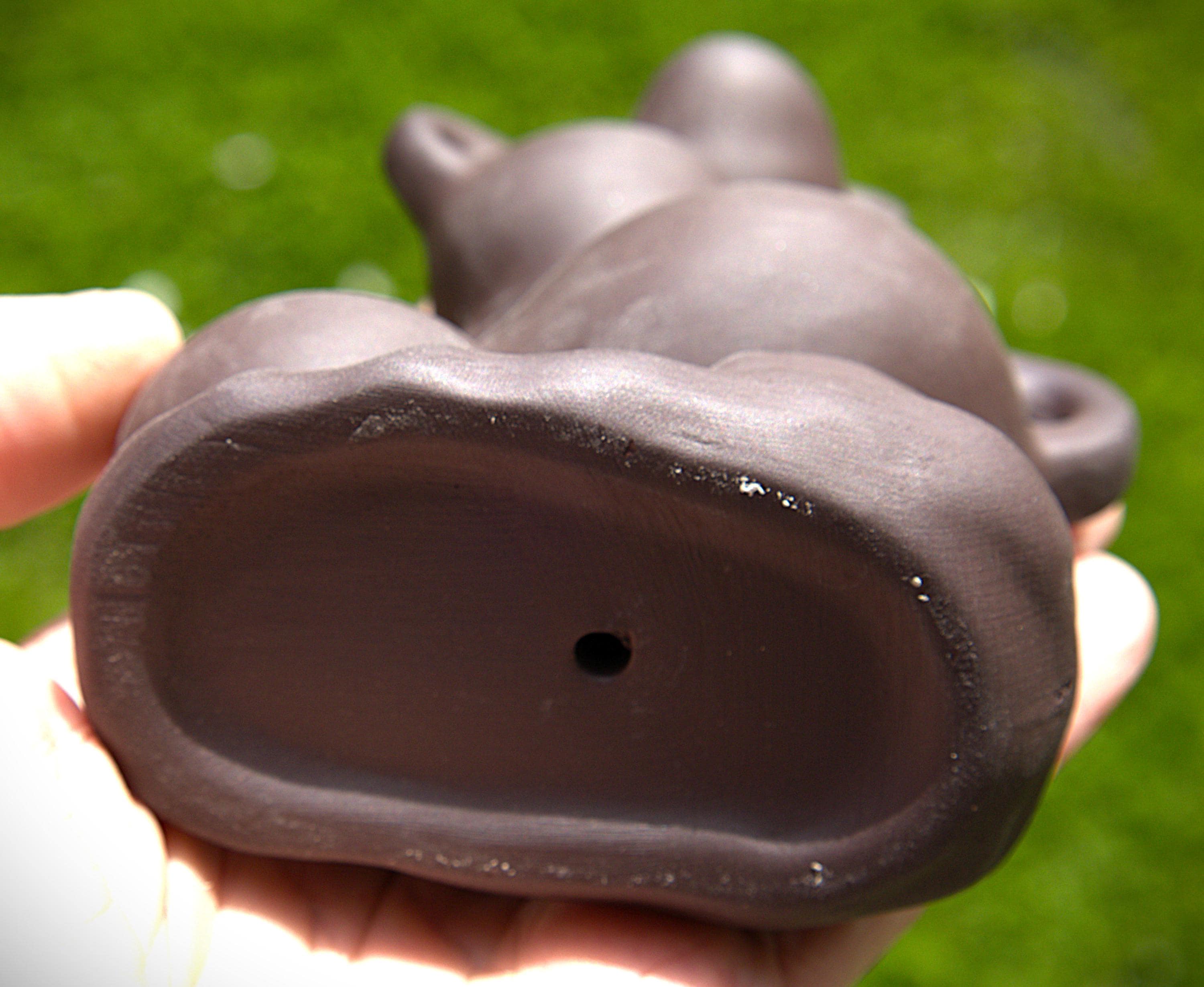 A close-up of a dark brown ceramic teapot being held in a hand, showing the bottom of the pot with a small drainage hole, against a blurred green grass background.