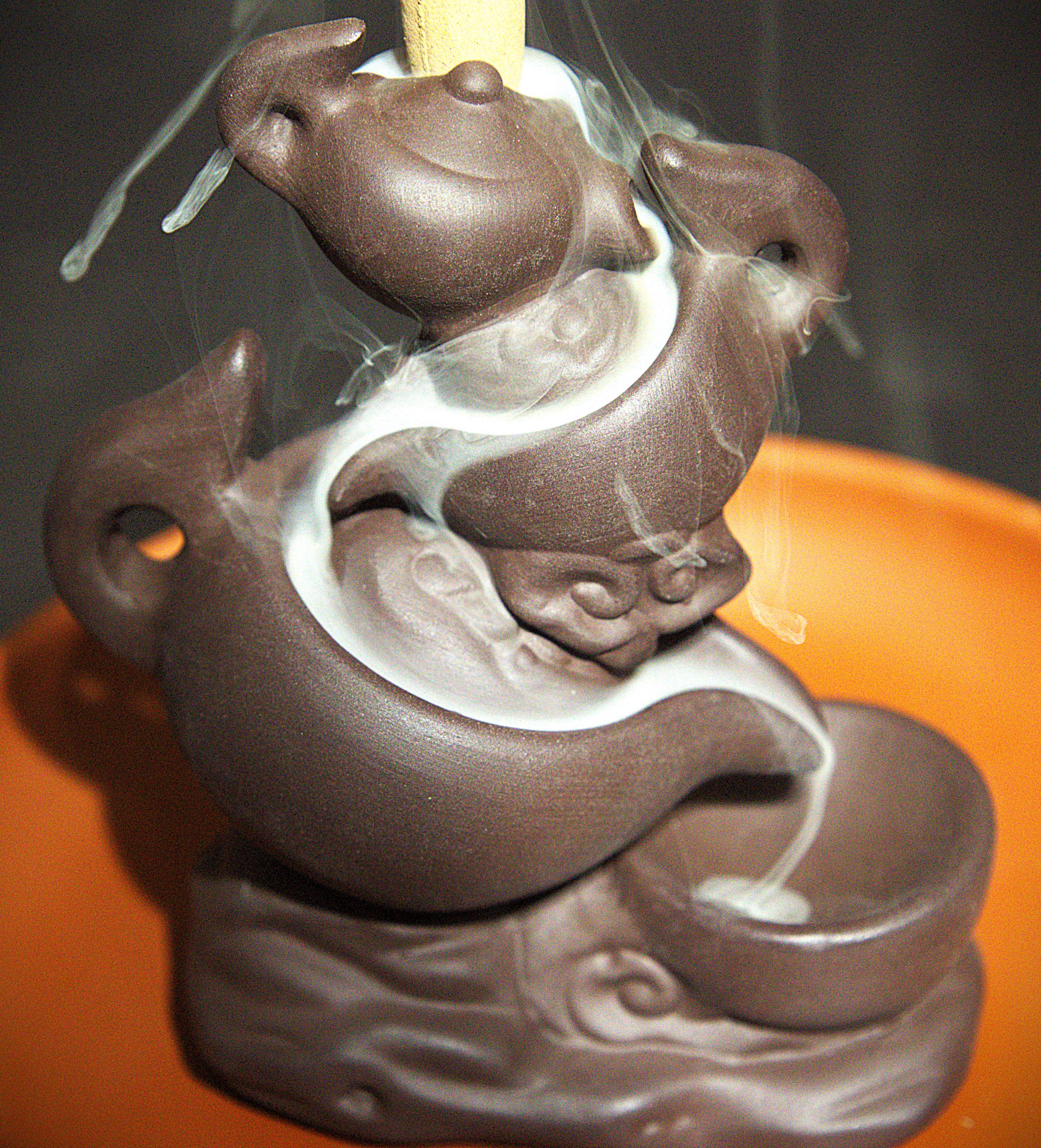 A close-up of a dark brown ceramic incense burner shaped like a stylized animal, with smoke rising from a small bowl at the top and white incense material flowing into a lower bowl, set against an orange surface.