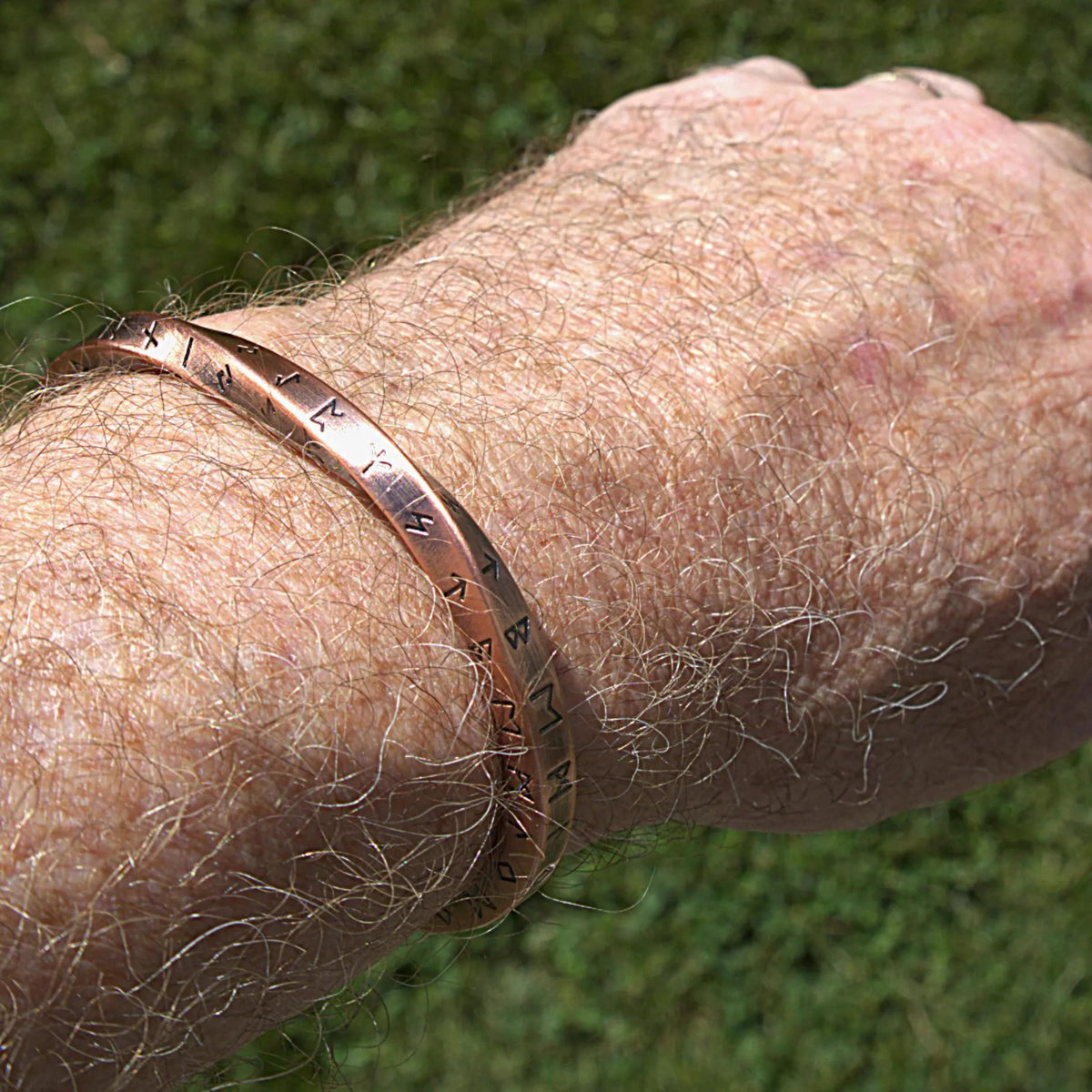 A close-up of a person's hairy forearm and hand wearing a copper-colored metal cuff bracelet engraved with runes, set against a blurred green grass background.