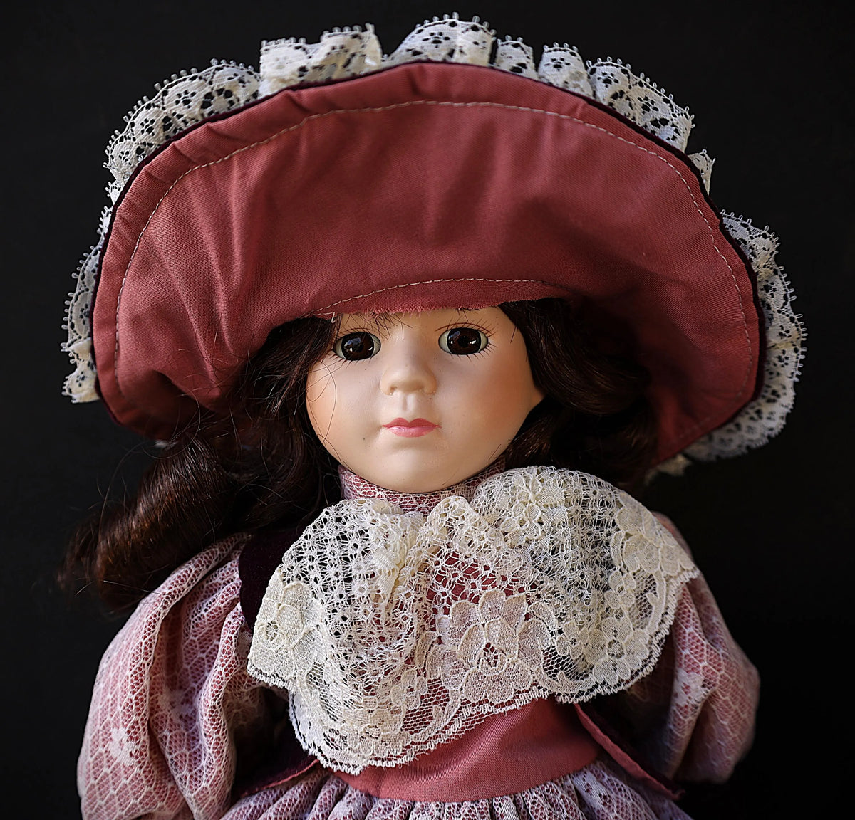 A vintage-style porcelain doll with dark brown hair, wearing a dusty rose satin bonnet with white lace trim and a matching lace collar, set against a solid black background.
