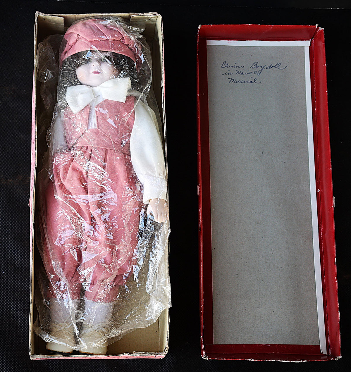 A vintage collectible porcelain doll in its original cardboard box, shown next to its red-lined lid which features a handwritten inscription. The doll is wrapped in protective plastic and wears a pink dress, matching bonnet, white bow, and white shirt.