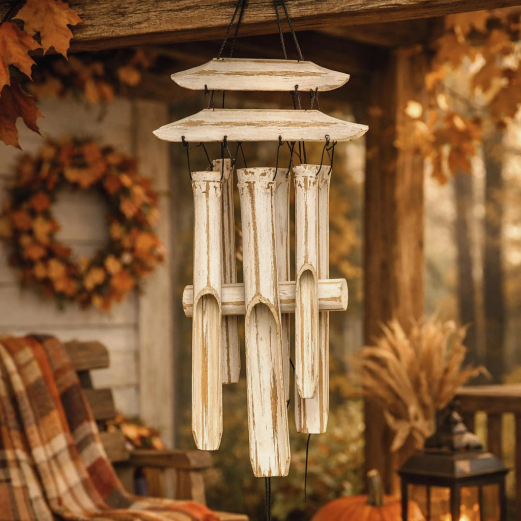 A rustic outdoor wind chime with layered horizontal plates and six vertical bamboo tubes, shown hanging on a porch with autumn decorations including a wreath, blanket, lantern, and pumpkins.