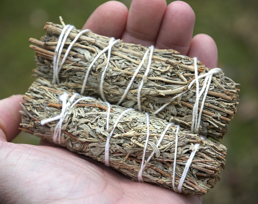 Two dried sage smudge bundles wrapped in white string, held in a person's hand against a blurred green outdoor background.